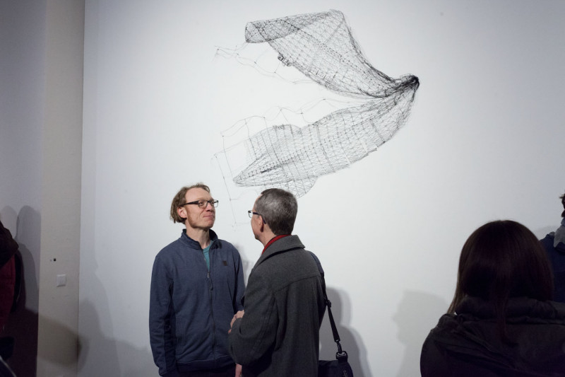 People are talking in front of a work of art. A fine wire sculpture hangs on the wall.