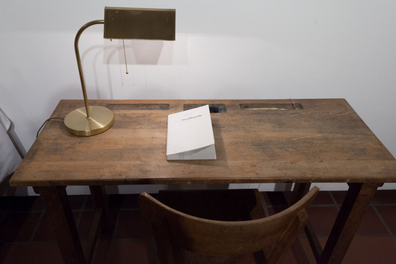 A wooden table with a chair and a lamp stands against the wall. On the table is a booklet with the inscription: Encyclopaedia.