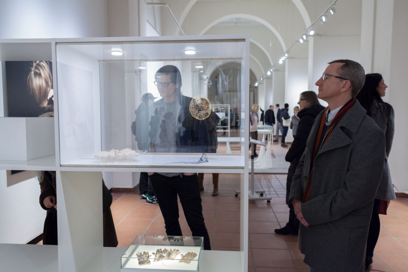 Ausstellungsansicht. Besucher betrachten die Kunstwerke. Es steht ein Vitrinenschrank, mit kleinen feinen Gegenständen darin, im Raum.