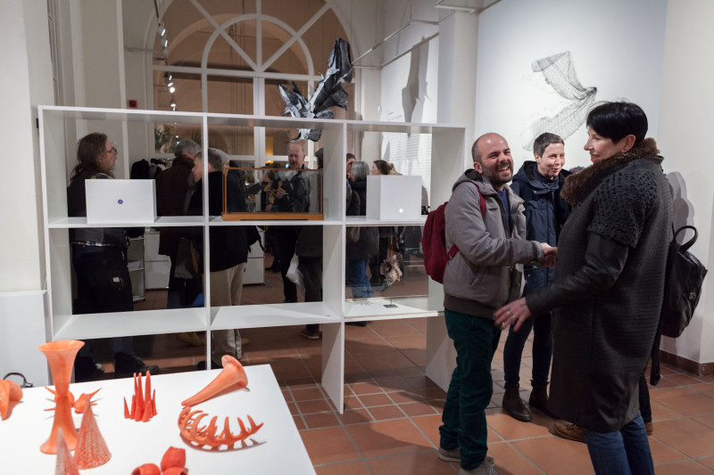 Besucher betrachten die Ausstellung. Im Vordergrund sind orangene Skulpturen ausgestellt.