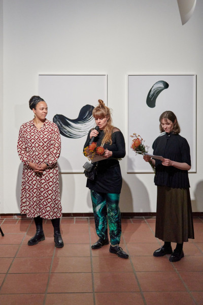 Drei Frauen stehen im Galerieraum, eine von ihnen spricht in ein Mikrofon