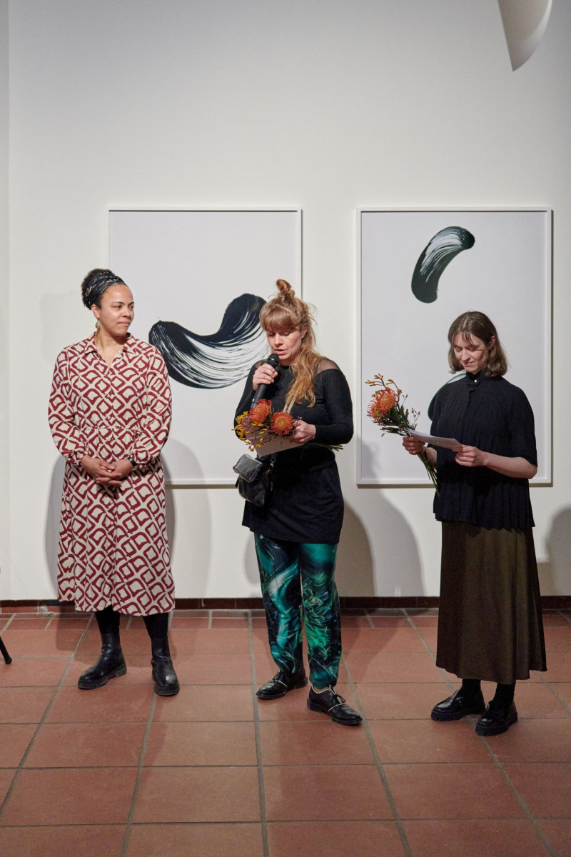 Three women are standing in the gallery space, one of them is speaking into a microphone