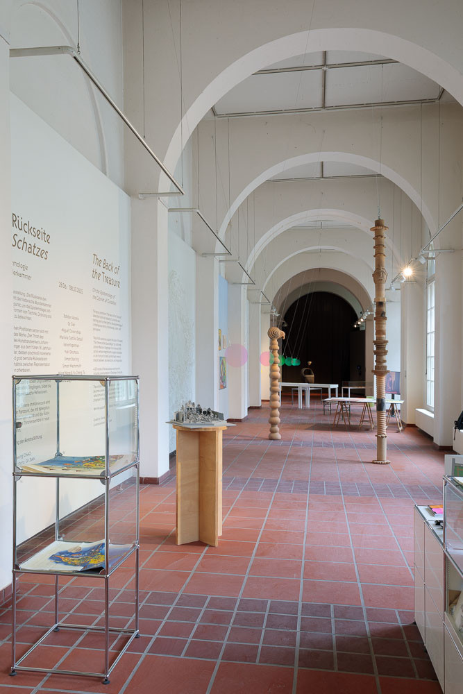 Galerieraum mit Rundbögen. Kunstwerke in Form von säulenartigen Skulpturen. Weitere Objekte in Vitrinen und auf Tischen präsentiert