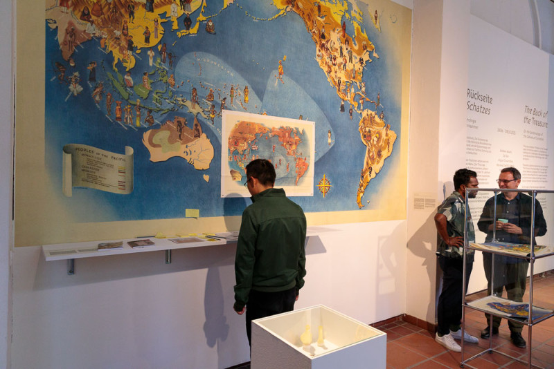 Ausstellungsbesucher vor und neben einer vom Künstler umgestalteten Weltkarte. Davor Vitrine mit Kunstobjekten