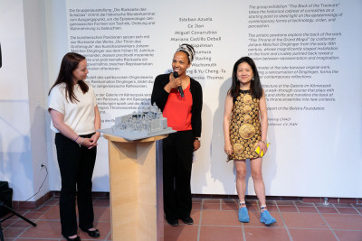 Three women standing in front of the introductory text on the wall. The gallery director stands next to the curator and speaks into the microphone