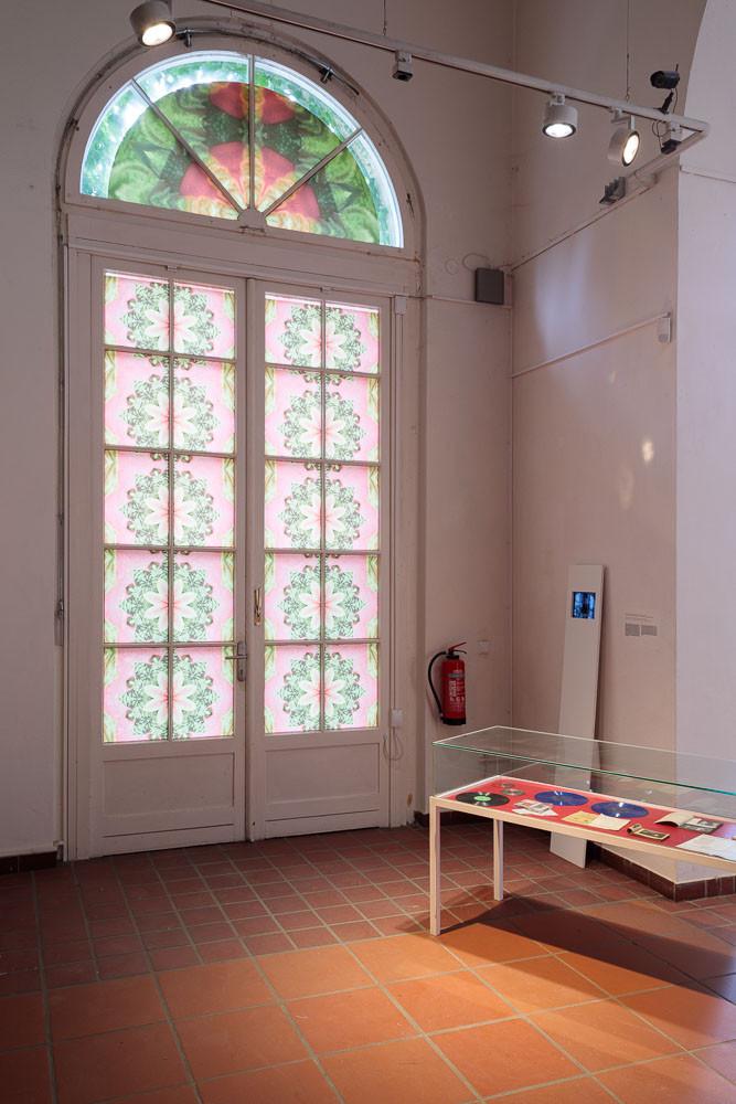 Tablet in einer Platte eingefasst an der Wand lehnend und Objekte wie Schallplatten und Hefte in einer Glasvitrine