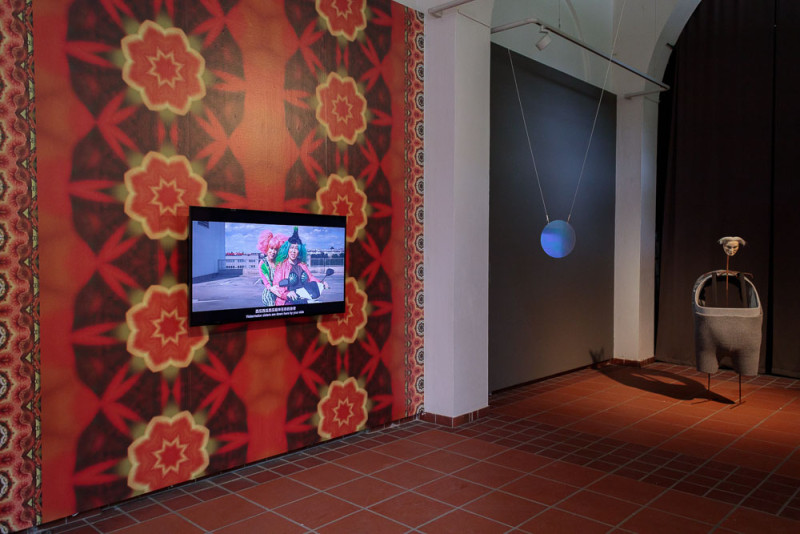 Monitor an der Wand mit Videoarbeit. Am rechten Bildrand eine stehende Installation mit Maske