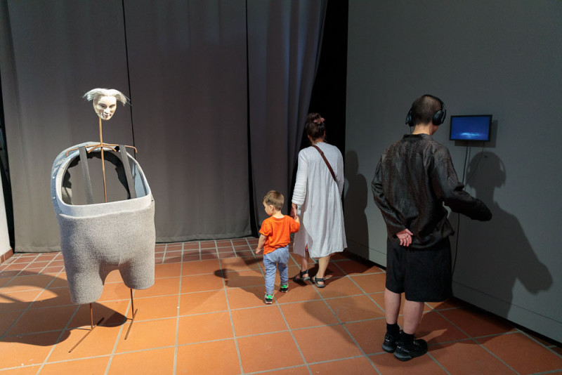 Stehende Installation mit Maske, Tablet mit Videoarbeit an der Wand, dazwischen Ausstellungsbesucher