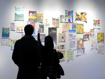 Vor einer weißen Wand mit verschiedenen bunten Drucken stehen, von hinten fotografiert,  ein Mann und eine Frau.