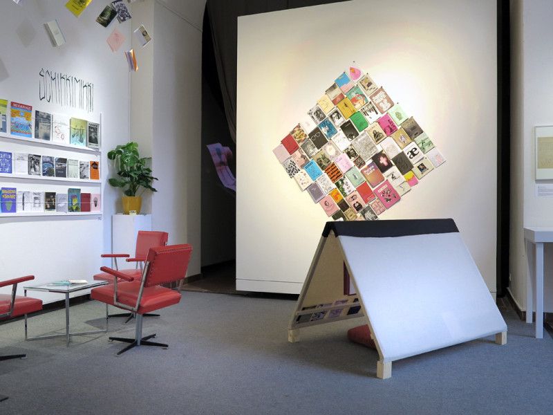 An der linken Seite stehen drei rote Drehsessel an einem kleinen Glastisch. Darüber sind bunte Broschüren in einem 3etagigen Regal. Von der Decke hängen ebenfalls Broschüren. Im Hintergrund an einer weißen Wand sind bunte Hefte in einer Raute angebracht. Rechts davor steht in Form eines Zeltes weißes Platten.