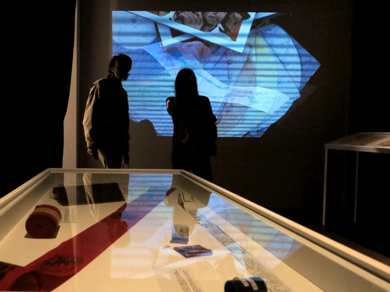 Im Vordergrund eine Glasvitrine  mit verschiedenen Gegenständen. Im Hintergrund stehen zwei Peronen vor einer Projektion,die digitalisierte Scans von Zines zeigt.