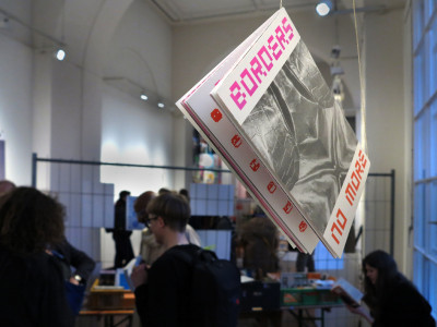 Im Vordergrund hängt von der Decke an einer Schnur eine Broschüre. Im Hintergrund etwas unscharf Gäste der Ausstellung.