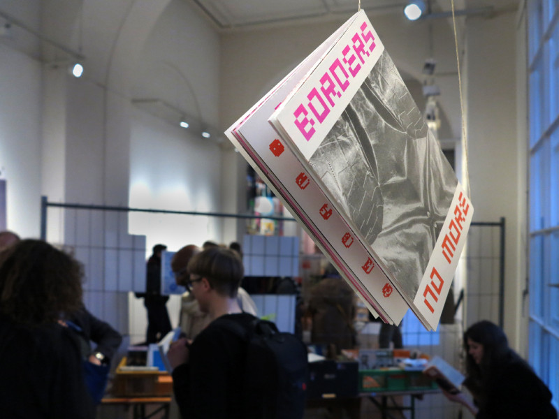 Im Vordergrund hängt von der Decke an einer Schnur eine Broschüre. Im Hintergrund etwas unscharf Gäste der Ausstellung.