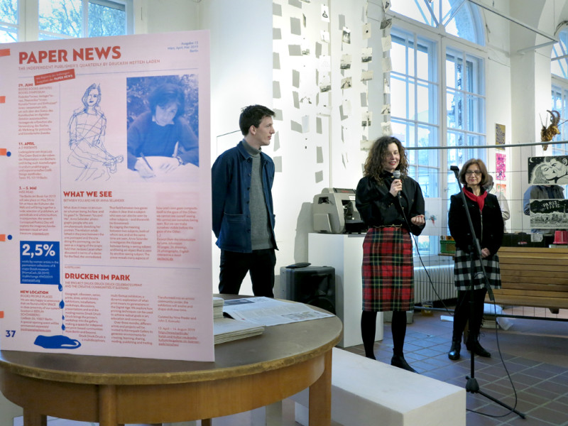 Blick in die Galerie.  Im Vordergrund links steht auf einem runden Tisch ein Plakat mit roter Schrift. Rechts daneben stehen die Kuratoren der Ausstellung mit einem Mikrofon in der Hand und die Galerieleiterin.