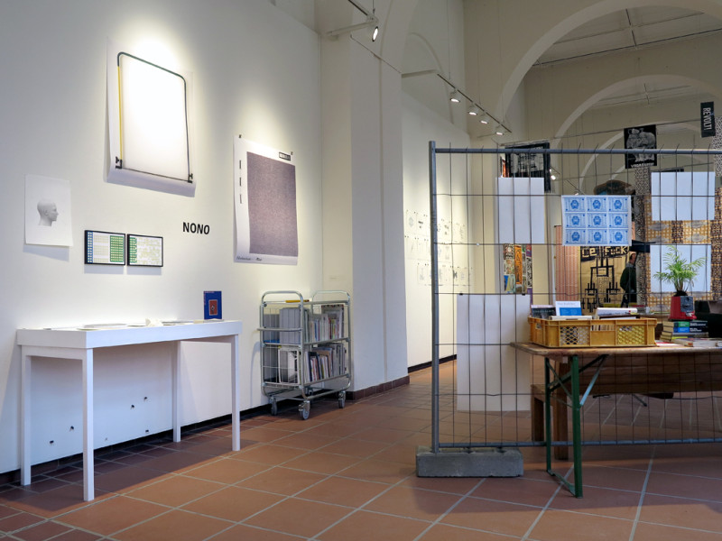 Mehrere schlichte Drucke hängen an der Wand. Davor ist die Bibliothek auf einer Vitrine und in einem Regal auf Rollen präsentiert.