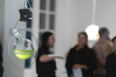 Detailed view of the artwork: A laboratory glass in the shape of a round bottom flask in a metal holder and connected with cables. Visitors stand in the background