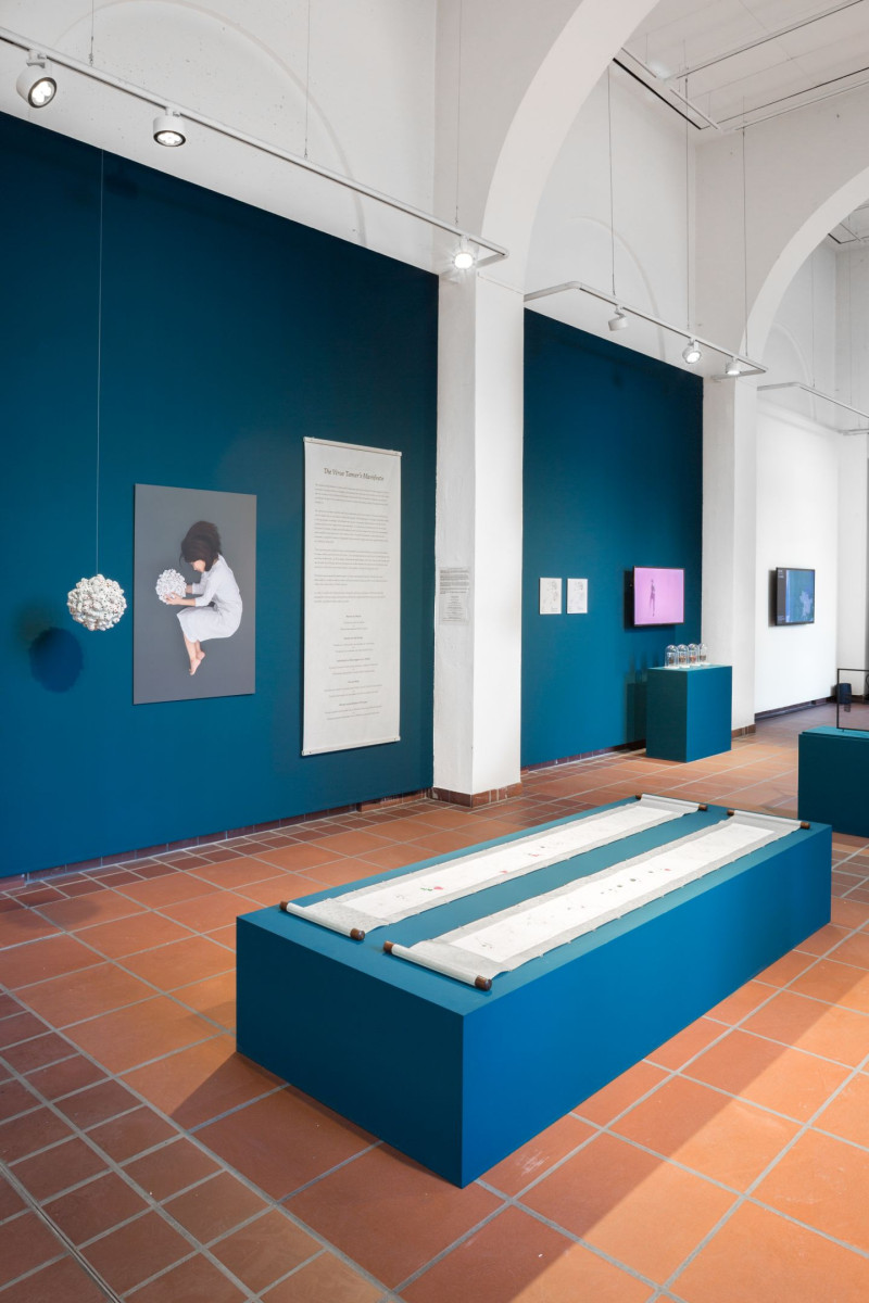 Ansicht des Galerieraums ohne Besucher. An der Wand sind Kunstwerke und Texte angebracht, darunter ein Bild, das eine kleine Person in einem hellen Gewand darstellt. Davor steht ein niedriges Podest, auf dem zwei ausgerollte Bildrollen liegen