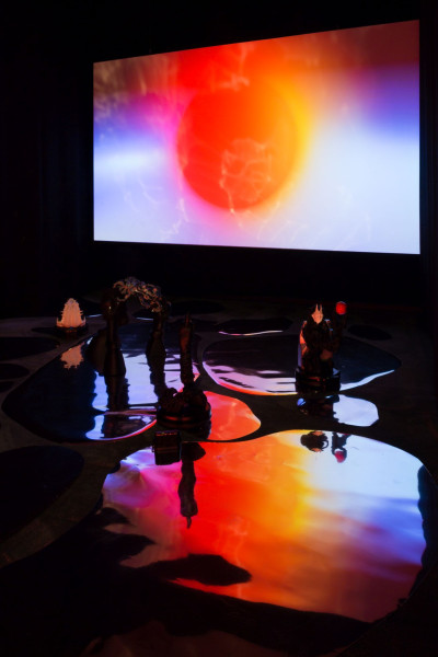 Large screen showing the artist's video work. In front of it, an installation with moving objects on mirror foils on the floor