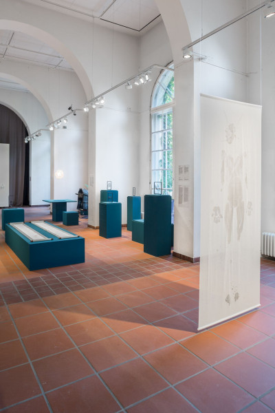 View of the gallery space. Works of art stand on pedestals of different heights and shapes. A work of art in the shape of a cotton cloth hangs vertically from the ceiling. The outline of a person is shown on the cloth