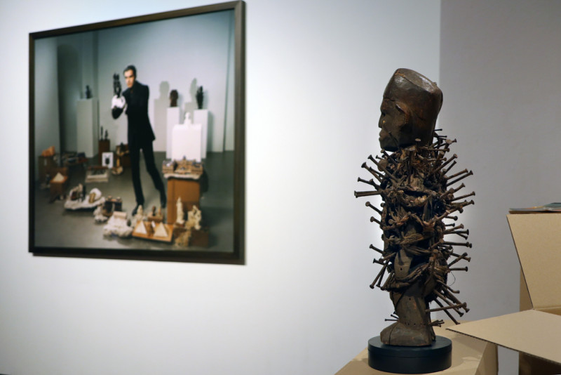 Eine Figur mit vielen Nägel in ihrem Körper im Vordergrund, im Hintergrund ein großformatiges Foto mit einem Mann umgeben von verschiedenen Objekten enthografischer Sammlungen.