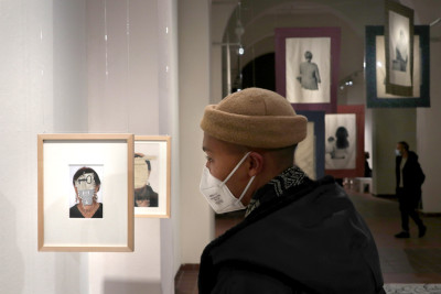 Eine Person schaut sich ein Bild an, das von der Decke hängt. In einem hellen Holzrahmen ist ein Fraunportrait zu sehen, das anstelle des Gesichts ein Versatzstück eines Dokuments zeigt.