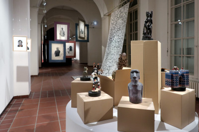 Im Vordergrund ist eine runde Bühne zu sehen auf der Kartons mit verschiedenen Objekten aus einer enthnologischen Sammlung stehen. Im Hinergrund die Ansicht des Ausstellungsraums.