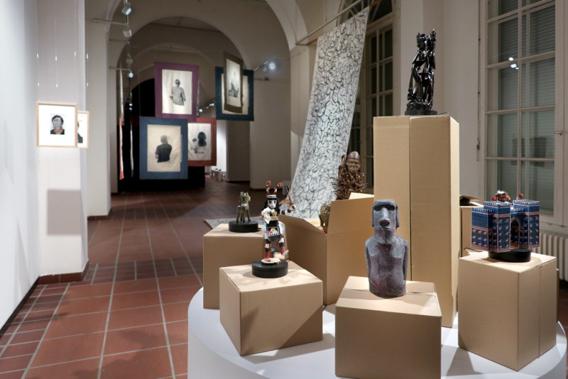 Im Vordergrund ist eine runde Bühne zu sehen auf der Kartons mit verschiedenen Objekten aus einer enthnologischen Sammlung stehen. Im Hinergrund die Ansicht des Ausstellungsraums.