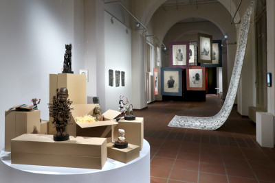 Im Vordergrund ist eine runde Bühne zu sehen auf der Kartons mit verschiedenen Objekten aus einer enthnologischen Sammlung stehen. Im Hinergrund die Ansicht des Ausstellungsraums.