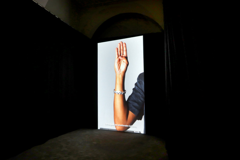 In einem dunklen Raum ist eine hochformatige Projektion zu sehen mit dem Ausschnit eines Arms und einer Hand.