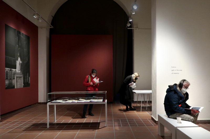 Ein Besucher mit Kopfhörer auf einer Bank rechts, links daneben eine Vitrine mit einer Besucherin dahinter vor einer roten Wand.