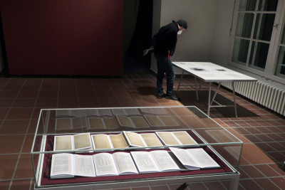 Eine Vitrine mit Büchern im Vordergrund, im Hintergrund eine rote Wand und ein Mann, der sich einen Tisch beugt.