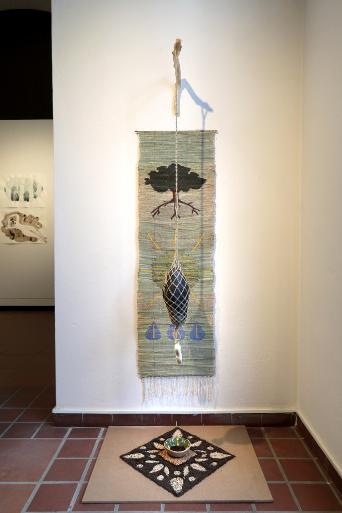 Various natural materials are woven into a hanging carpet and in front of it stands a vessel in the midst of an ornamental pattern made of natural materials.