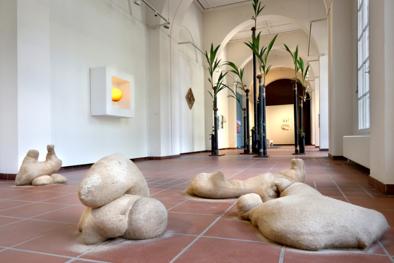 Im Vordergrund liegen amorphe Sandskulpturen auf dem Boden verteilt. Dahinter erheben sich mehrere in die Höhe ragende schmale Stelen mit kleinen Kokospalmen darauf.