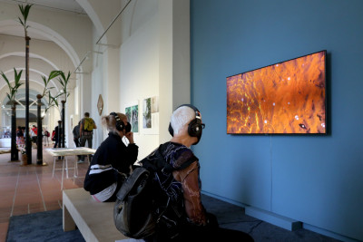 Visitors watch the film by Kari Roover. The screen shows a close-up of the sandy bottom of water.