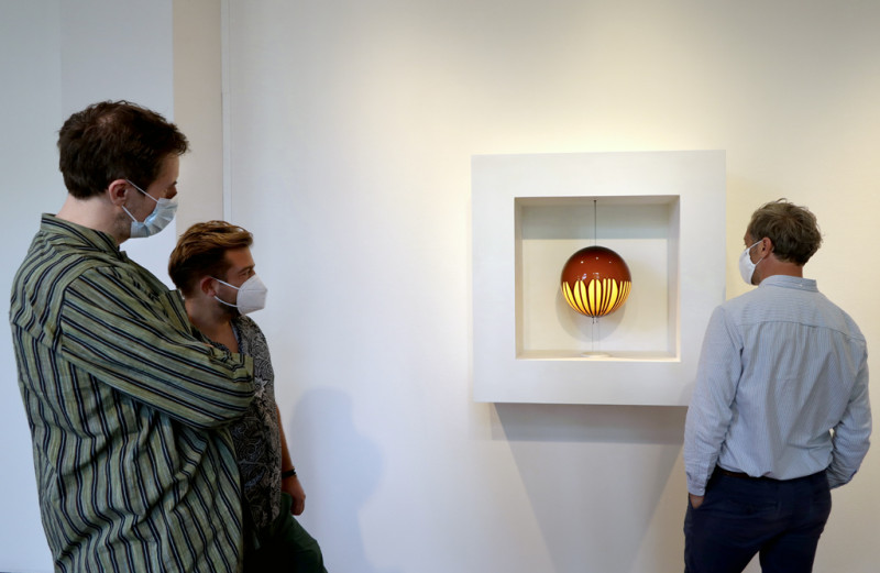 Visitors stand in front of a work that shows a glowing yellow sphere in a square white frame that is doused with brown liquid.