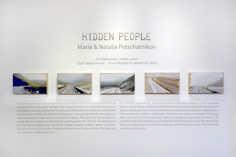 fünf Bilder hängen an der Wand, auf denen eine Straße zu sehen ist. Um die Bilder herum steht der Wandtext zur Ausstellung