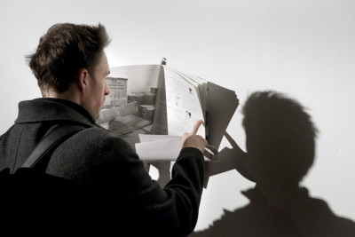 A man flips through the magazine TIEMPO MUERTO (Dead Time), which is mounted on the wall for viewing. He is pictured from behind