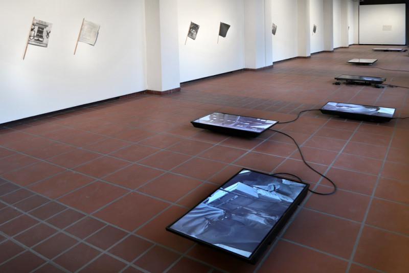 Ansicht des Galerieraums. Auf dem Boden sind mehrere Bildschirme verteilt. An den Wänden sind Exemplare der Zeitschrift TIEMPO MUERTO (Tote Zeit) befestigt