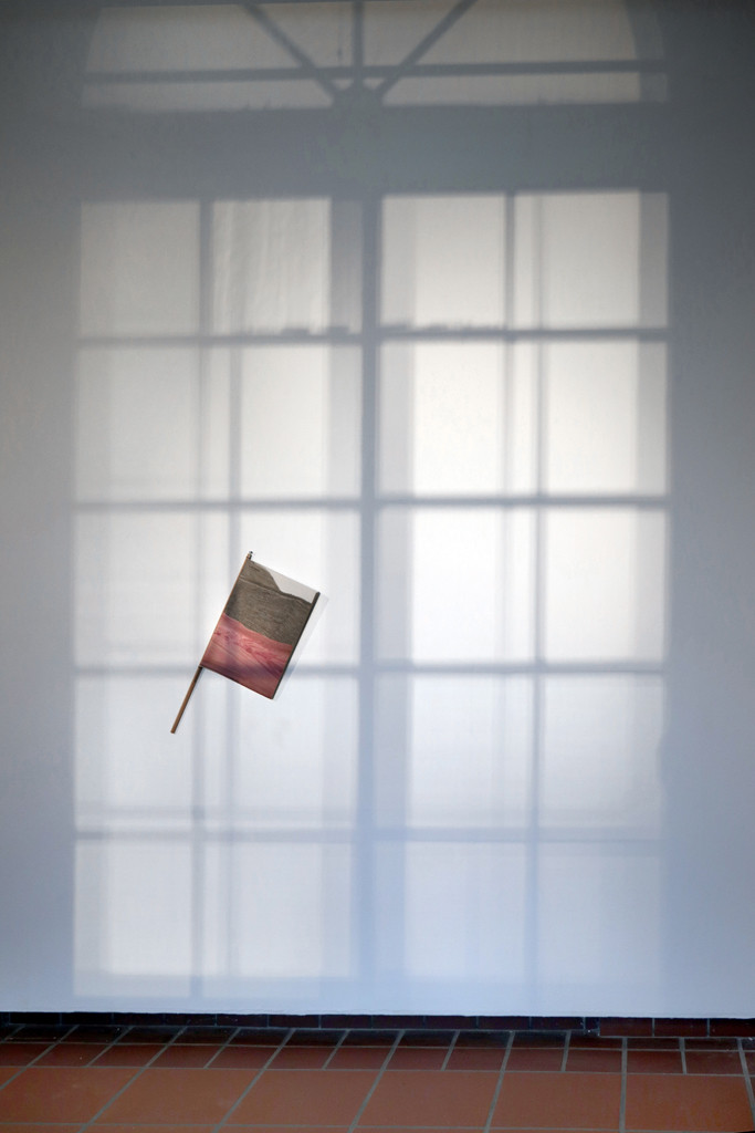 Zeitschrift TIEMPO MUERTO (Tote Zeit) um einen Stab gebunden und an der Wand zur Ansicht befestigt. Ein großer Teil der Wand ist mit einem Schatten in Form eines Rundbogenfensters abgebildet