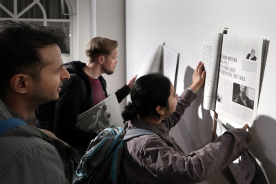Two men and a woman are looking at the magazine TIEMPO MUERTO (Dead Time), which is tied around a stick on the wall for viewing. Two of the people open the pages of the magazine