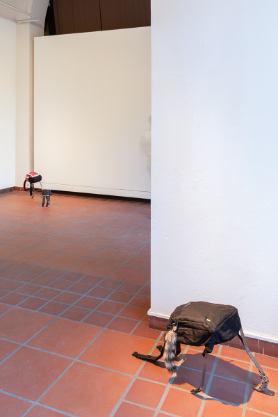 Two sculptural beings made from found materials occupy a terracotta-tiled gallery floor, with a large cream-colored wall panel in the background.