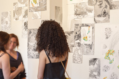 Two visitors viewing a gallery wall covered with mixed media artworks including textured paper pieces, ink drawings, and colorful illustrations arranged in an overlapping salon-style display