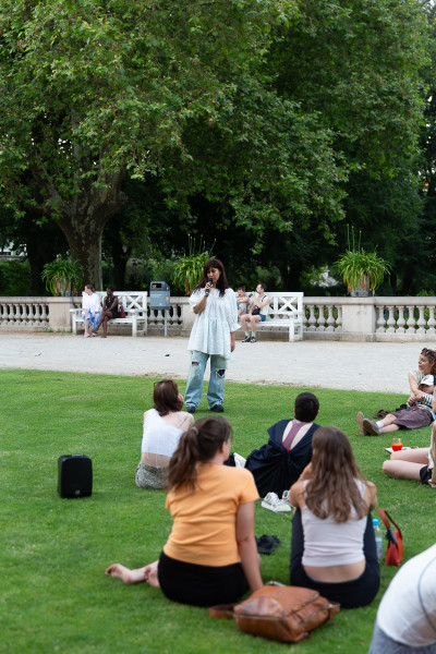 A woman stands speaking to a seated audience on a grass lawn beneath large trees in an urban park setting