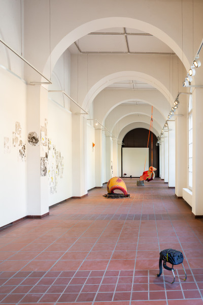 A vaulted exhibition hall with a brick floor, artwork hanging on the walls, and red and yellow installations on the floor