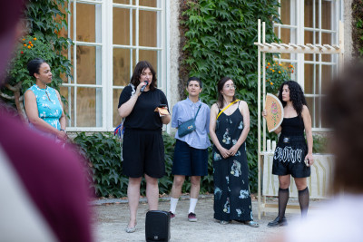 Five women stand in a courtyard with ivy-covered walls, one speaking into a microphone during a public event or presentation