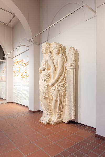 Cream-colored stone relief sculpture depicting two draped figures alongside architectural elements, displayed in a gallery corridor with informational panels.