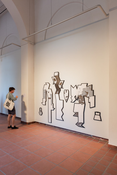 A visitor views an abstract line drawing mural on a white gallery wall depicting stylized figures with geometric forms and organic shapes in black ink.