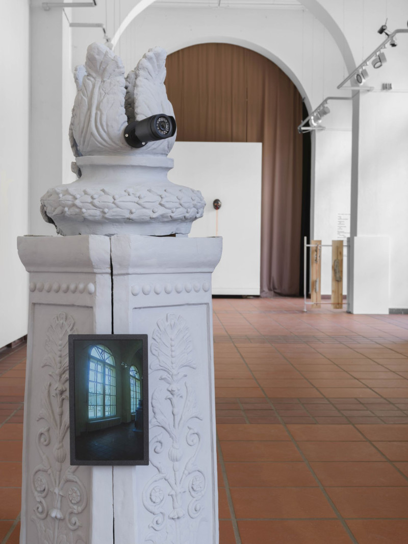 Ornamental verzierter Sockel, hergestellt als Gipsabguss eines Laternenpfahls in Minsk. Blätterförmiger Aufsatz, an dem eine Kamera angebracht ist. Am Sockelschaft ist senkrecht ein kleiner Bildschirm befestigt