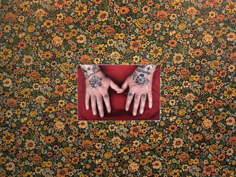 Blumentapete mit einem Foto im Mittelpunkt. Darauf sind alte tattoowierte Hände zu sehen.