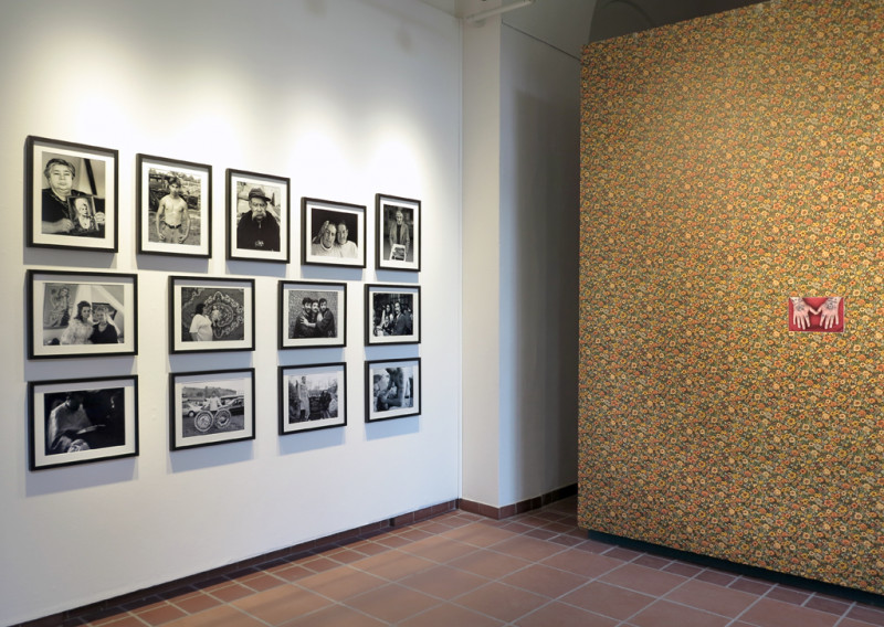 Ausstellungsansicht. An der Wand hängen eingerahmte schwarz-weiß Fotos. Daneben steht eine Wand mit Blumentapete und einem Foto in der Mitte.